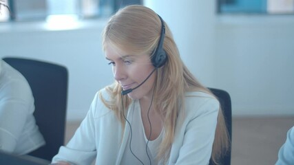 Serious fair haired operator in headset taking call, speaking at microphone at her workplace in call center. Static shot. Customers assistance or telemarketing concept