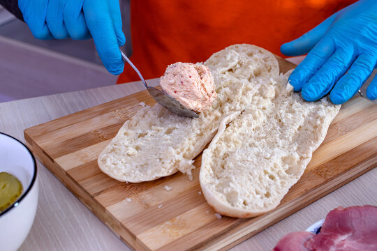 Chef preparing tasty fresh baguette submarine sandwich. Close up of hands in gloves spreading cream cheese with spicy red pepper, called urnebes, on a baguette sliced in half on wooden cutting board. 