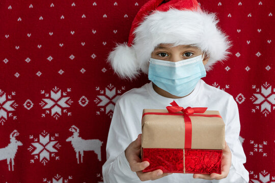 close up image of boy in santa hat and face medical mask holds two gift boxes tired with ribbons on red christmas background. Holiday concept