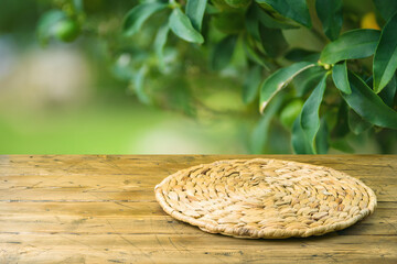 Obraz premium Empty wooden table with wicker round placemat over green garden blurred background. Spring mock up for design
