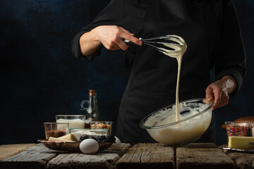 The chef kneads the dough with a whisk in glass bowl. Backstage of cooking waffle on wooden table with ingredients on dark blue background. Frozen motion. Traditional recipe.