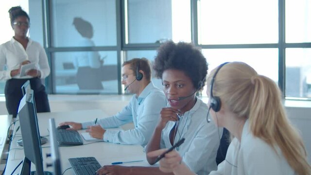 Female mentor watching new call center operators in headsets working at computers, doing tasks, consulting each others. Female business leader or telemarketing concept