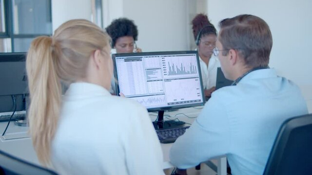 New employee asking mentor about financial data on monitor. Diverse team of financial brokers in headsets. Back view. Finance or trading concept