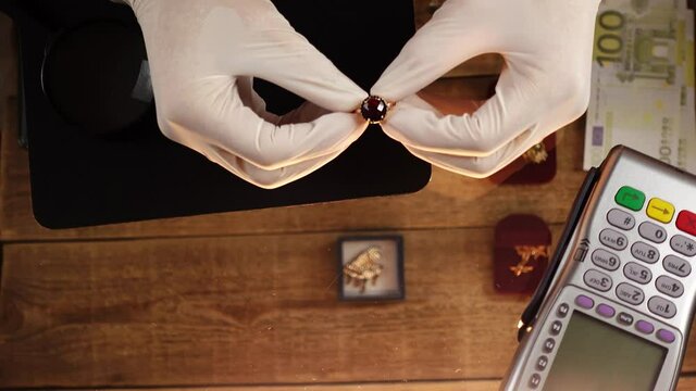 Close-up of hands in gloves examinating a gold ring with a gem. An owner of a pawnshop evaluates attentively jewellery sitting at his transparent glass table. Concept of buying up of precious metals.