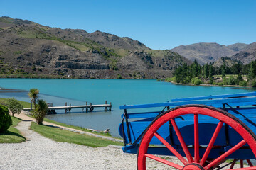 Fototapeta premium Red and blue cart at top of path leading to jetty into turquoise river surrounded by Central Otago hills at Cromwell historic precinct