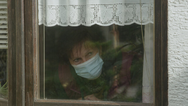 CLOSE UP: Depressed Elderly Woman Wearing A Facemask Looks Out Her Window.