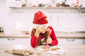 Little teenage girl sits in the kitchen and holds her head, painting gingerbread cookies with icing.