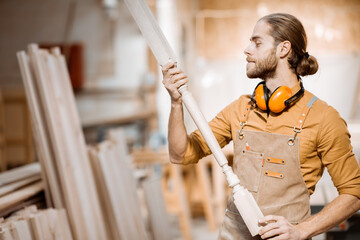 Handsome carpenter in uniform working with wood, checking the quality of the wooden baluster at the joinery