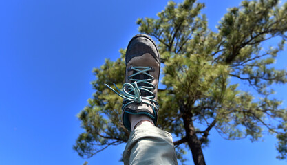 A person is raising his or her a right leg with shoe to blue sky with a blur pine tree in background