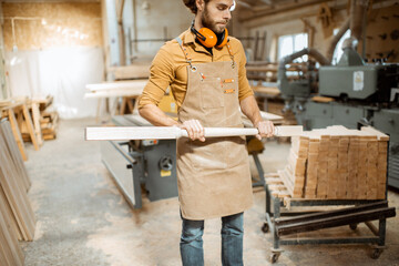 Handsome carpenter in uniform working with wood, checking the quality of the wooden baluster at the...