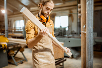 Handsome carpenter in uniform working with wood, checking the quality of the wooden baluster at the joinery