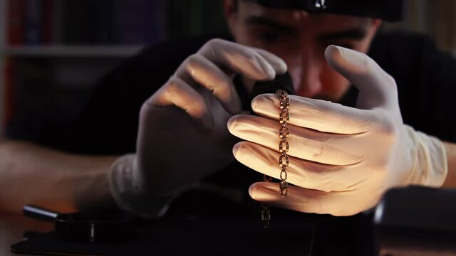 A man appraises gold bracelet with the help of magnifying glass in close-up. Portrait of working goldsmith who looking at chainlet through loupe. Concept of pawnshop business and buying precious metal