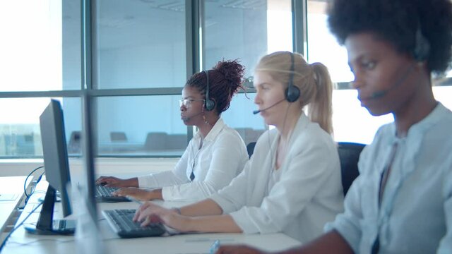 Focused diverse operators in headsets working at computers in call center, sitting in row and typing. Side view. Contact center or telemarketing concept