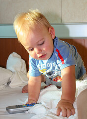 Un niño pequeño rubio juega encima de la cama con un telecomando. Retrato con luz natural