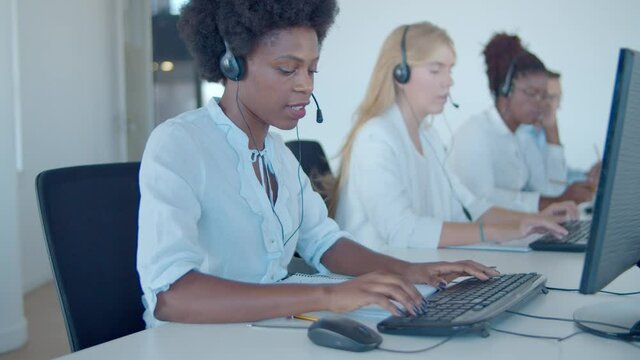 Team of call center operators in headsets sitting in row, using computers while speaking at microphones. Customers assistance or telemarketing concept.