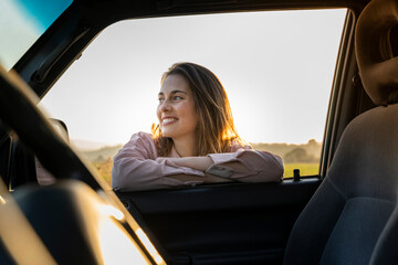 Happy young woman looking away while leaning on car window during sunset