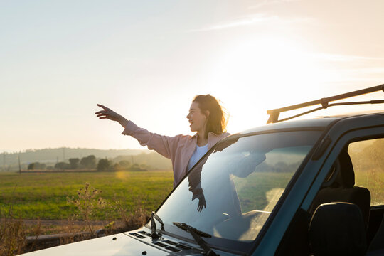 Cheerful woman leaning out of car during road trip against sky