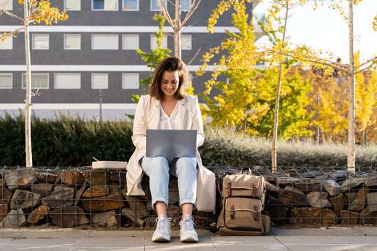 Smiling businesswoman using laptop while sitting on retaining wall in city