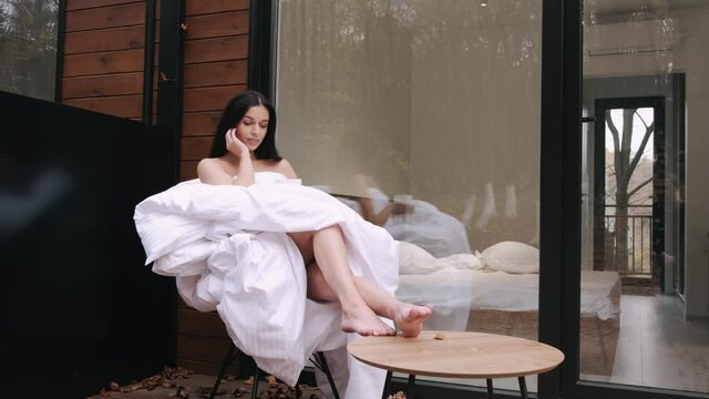 Young brunnete woman relaxing in forest house. The naked lady is wrapped in blanket and having a cup of coffee enjoying time alone.
