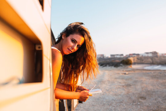 Woman with book and pen looking out of camper van door at beach