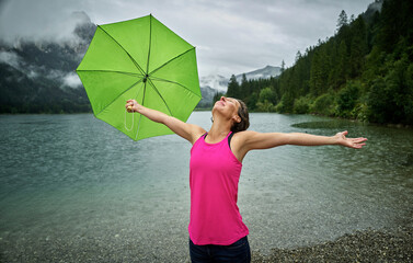 Happy woman holding umbrella while standing with arms outstretched at Haldensee lakeshore during rainy season