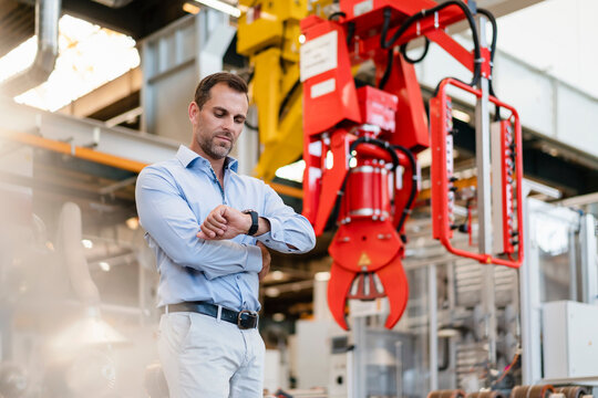 Male entrepreneur checking time on wrist watch while standing by claw at factory