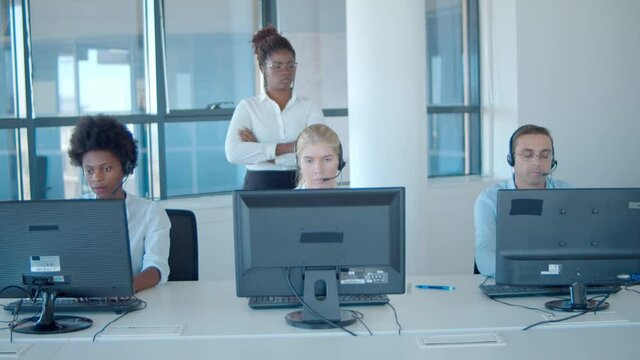 Strict female boss watching call center employees in headsets working at computers. Front view. Female business leader or telemarketing concept