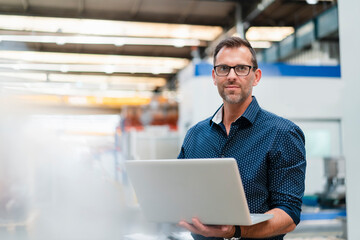 Smiling businessman holding laptop while standing in industry
