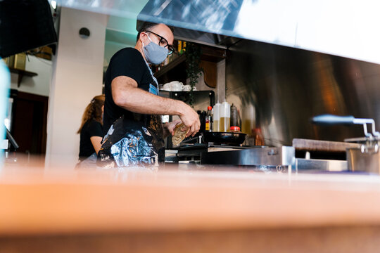 Male barista wearing protective face mask while cooking in kitchen of coffee bar during COVID-19