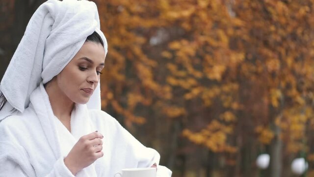Young woman on the balcony holding a cup of coffee in the morning. She is in hotel room looking at the nature in autumn. Her hair is wrapped in the bath towel on her head and she is wearing bath coat.