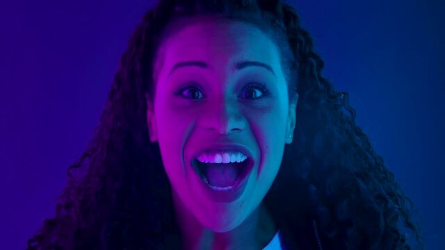 Portrait of a young lovely female African American looking at the camera with shocked and surprised wow face expression. Close up face illuminated with purple and blue neon lights. Slow motion.
