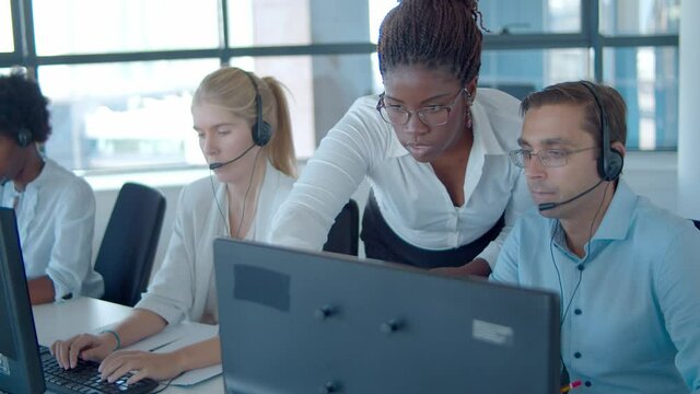 Serious African American manager helping call center workers with new software, pointing at monitors and speaking. Customers assistance or telemarketing concept.