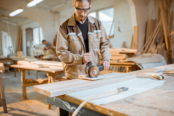 Handsome carpenter in uniform brushing wood with hand machine at the carpentry manufacturing