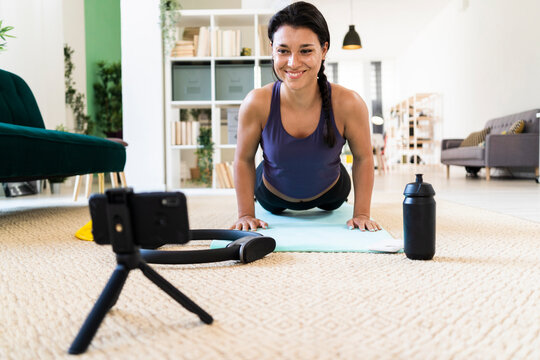 Smiling young woman video recording on camera while doing push ups at home
