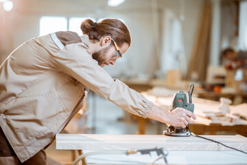 Handsome carpenter in uniform chamfers wooden bar with a hand machine at the carpentry manufacturing