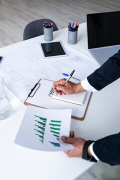 Hands of male entrepreneur analyzing growth graph while writing in diary at creative workplace