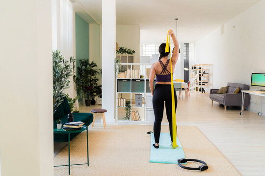 Young woman stretching with resistance band standing at home - Powered by Adobe