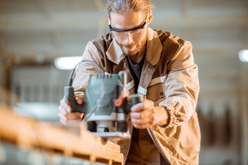 Handsome carpenter in uniform chamfers wooden bar with a hand machine at the carpentry manufacturing