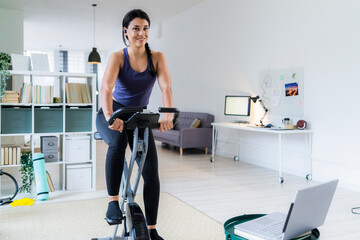 Smiling young woman listening music through bluetooth while sitting on exercise bike at home