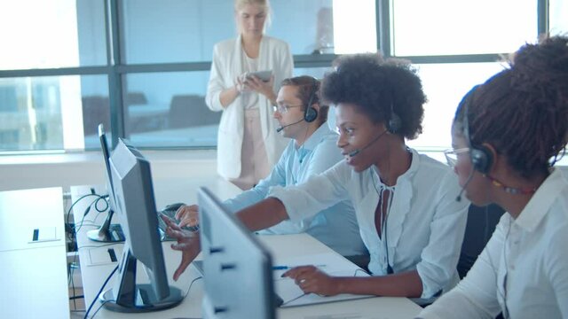 New call center operators in headsets working at computers and consulting each others while female manager standing by them and using tablet. Female business leader or telemarketing concept