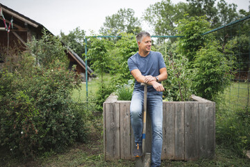 Smiling handsome man holding spade looking away while leaning on wooden raised bed at vegetable garden