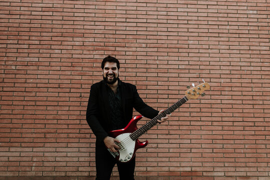 Cheerful musician playing guitar while standing against brick wall