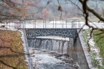 Obraz premium A lake embankment with outlet, a stone bridge with railing. White snow on green grass. Branch with dry leaves framing the image.