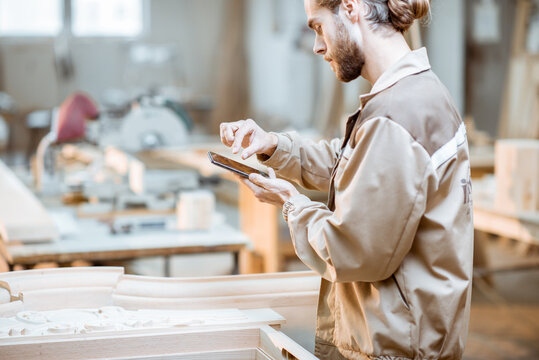 Handsome carpenter working on wooden carvings using a smart phone at the carpentry manufacturing. Concept of using mobile technologies in carpentry