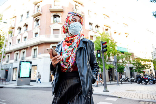 Muslim woman in hijab holding mobile phone walking on street in city during COVID-19