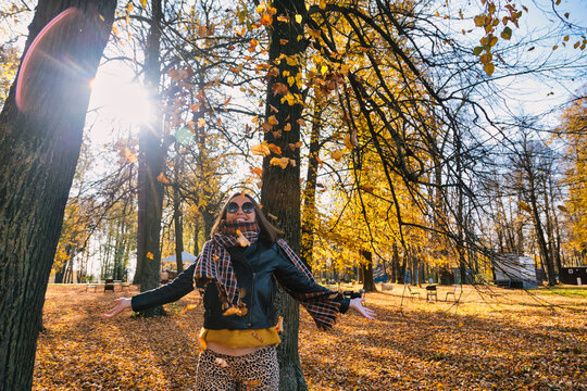 Happy woman standing with arms outstretched under falling dry autumn leaves while against trees at park