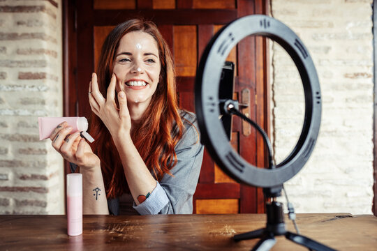 Smiling woman applying cream while filming make up tutorial on mobile phone at home