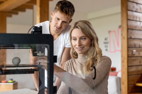 Businesswoman operating 3D printer while sitting at home by colleague standing behind