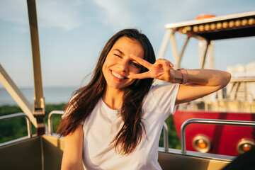 Smiling beautiful woman showing peace sign and winking while enjoying Ferris wheel ride