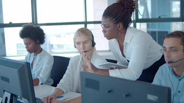 Young African American manager helping call center employees with new software, pointing at monitors and speaking. Customers assistance or telemarketing concept.